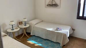 A small, neatly arranged bedroom with a single bed, a small table with a lamp, a framed picture on the wall, and a window. - Apartment in Porto Pollo Near Surf Paradise (Palau)