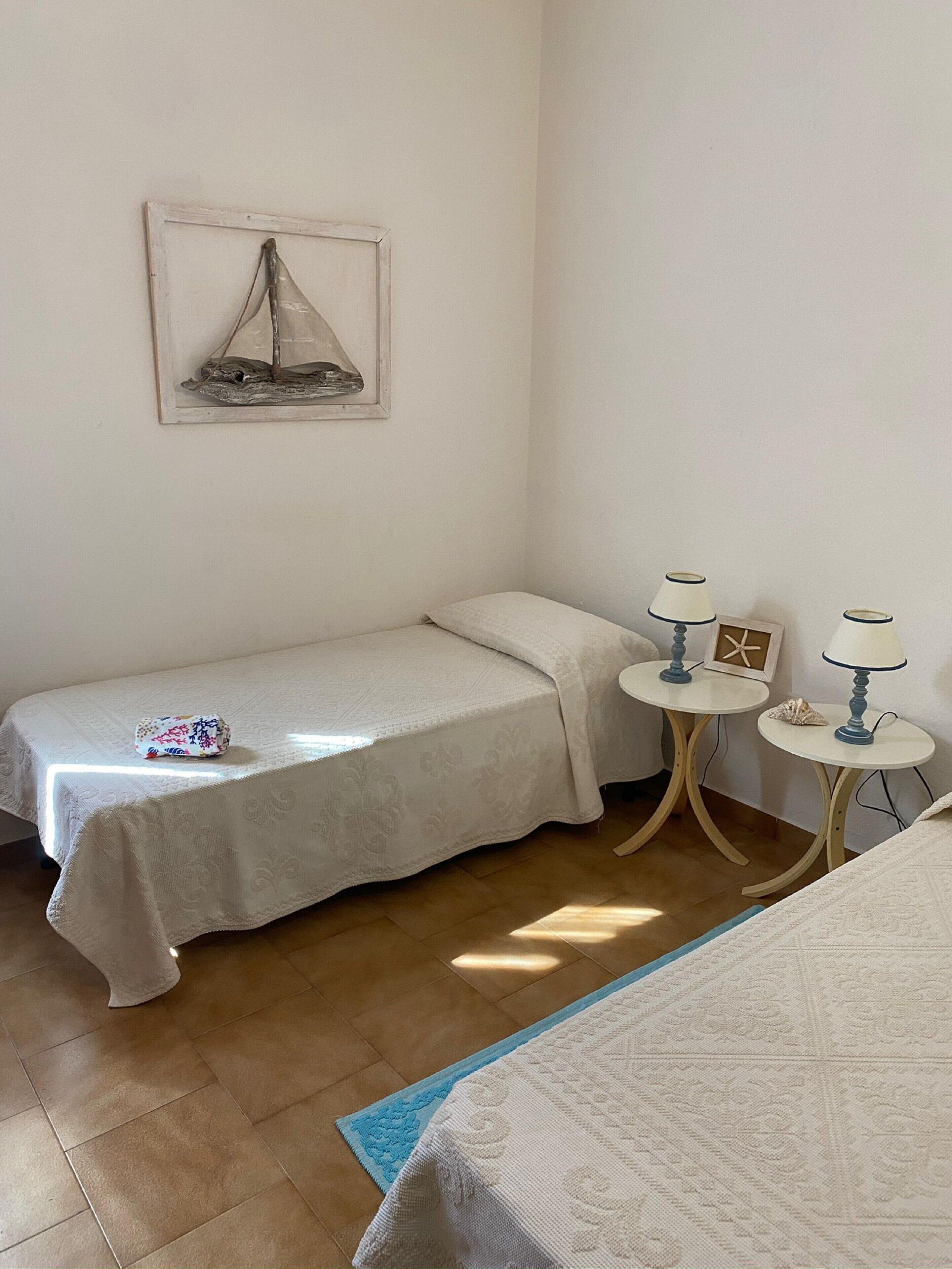 Quarto pequeno e bem arrumado com duas camas de solteiro, uma mesa de cabeceira com abajur, uma pequena mesa com abajur e uma moldura com imagem na parede.