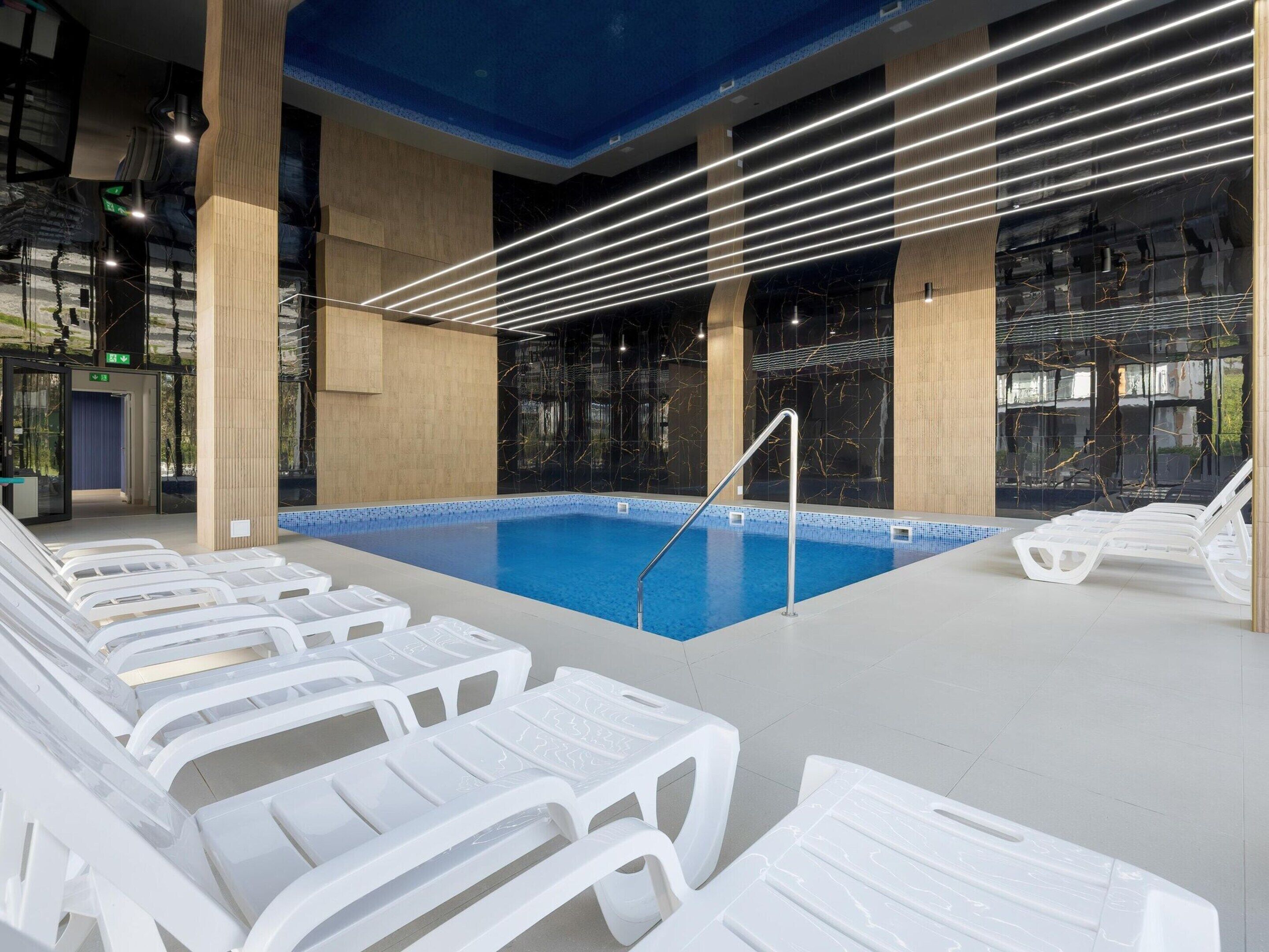 Ein Hallenbad mit klarem, blauem Wasser, umgeben von weißen Liegestühlen und einem modernen architektonischen Design mit blauer Decke und Glaswänden.