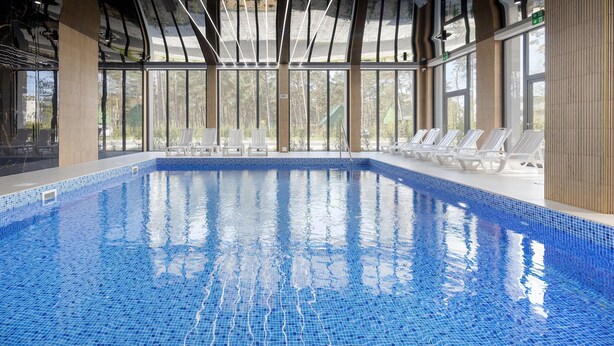 Ein überdachter Pool mit großen Glasscheiben, weißen Liegestühlen und einem gefliesten Boden.