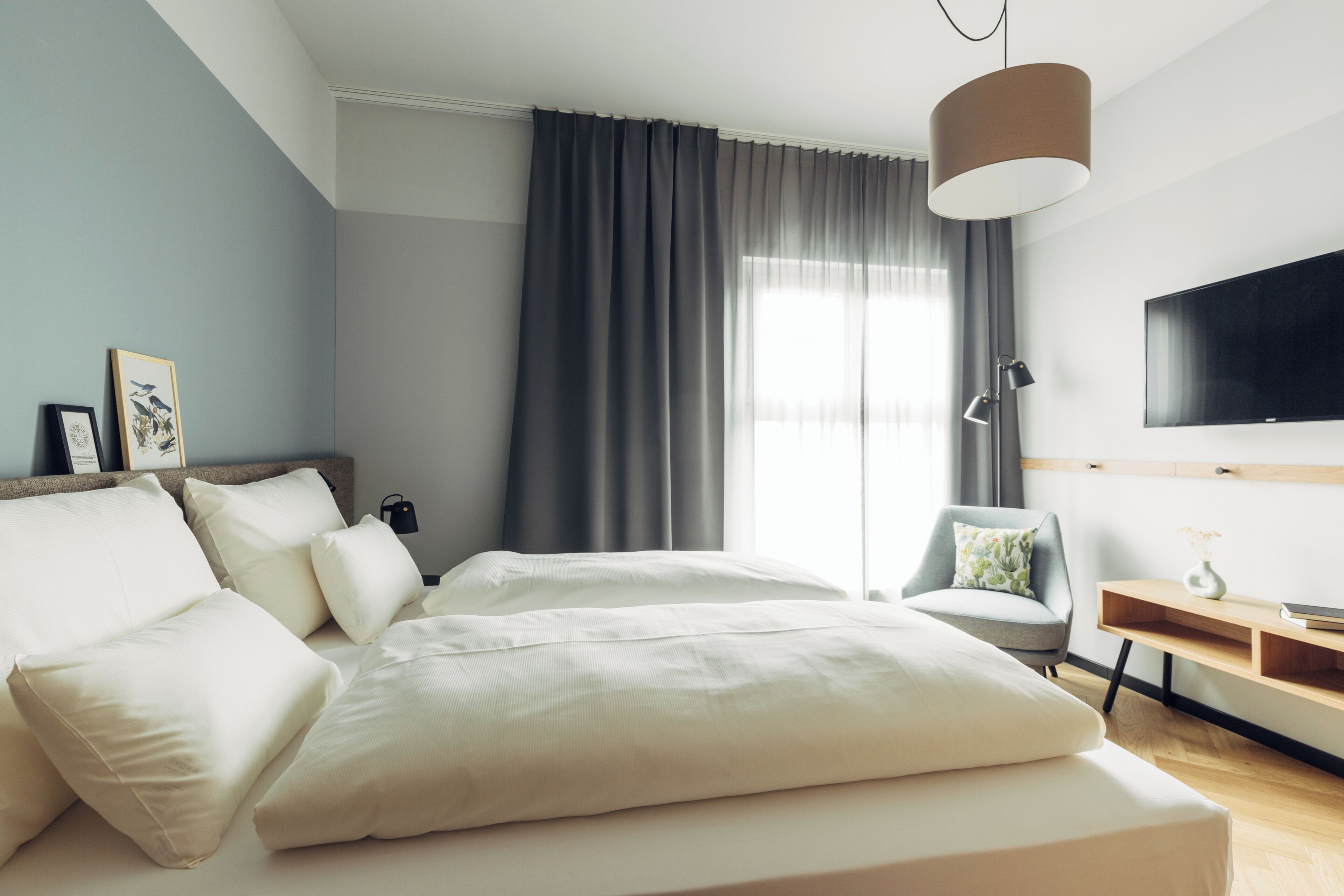 A modern hotel room with a large bed, a desk, a chair, a TV, and a window with curtains.