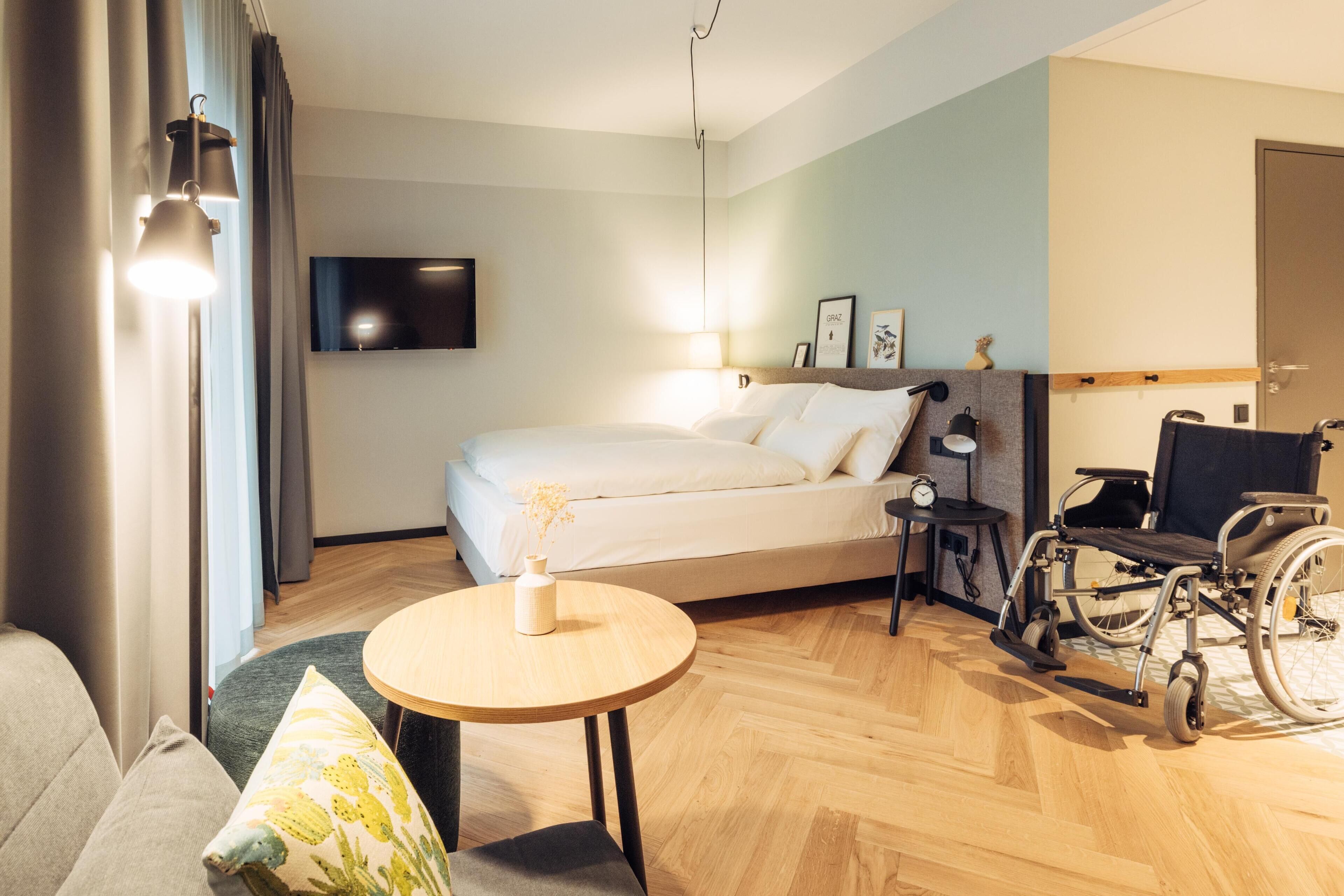 A hotel room with a bed, wheelchair, bedside table, lamp, and a TV mounted on the wall.
