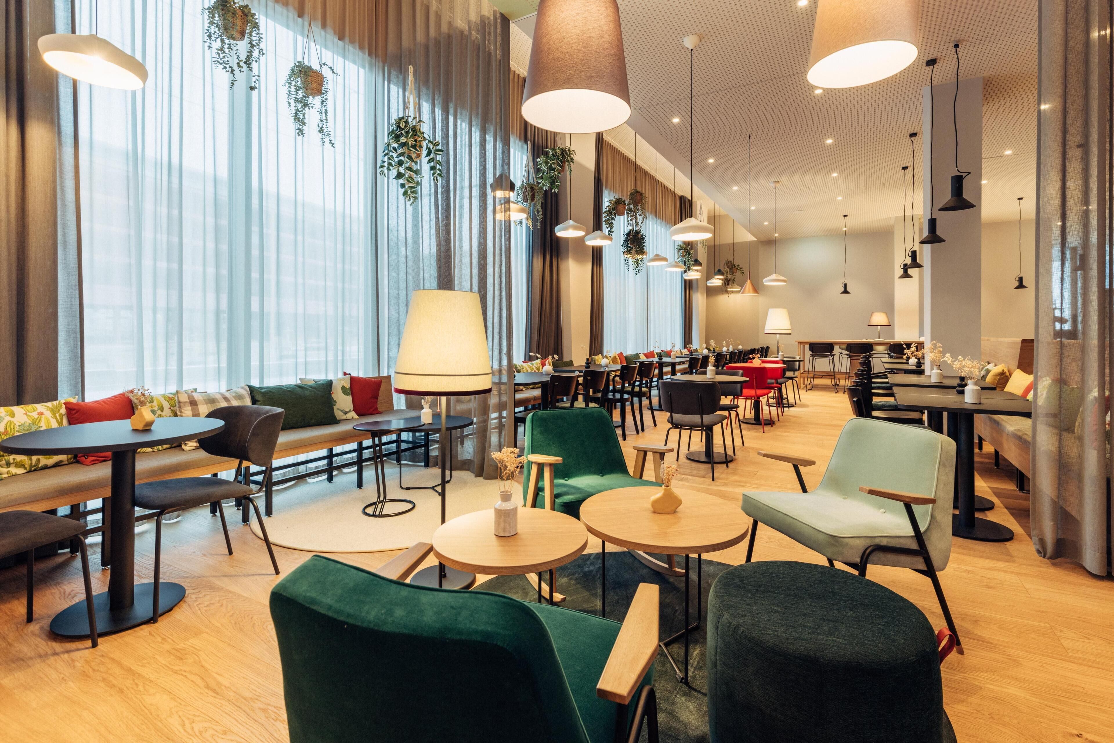 A modern dining area with round tables, green upholstered seating, and hanging plants.
