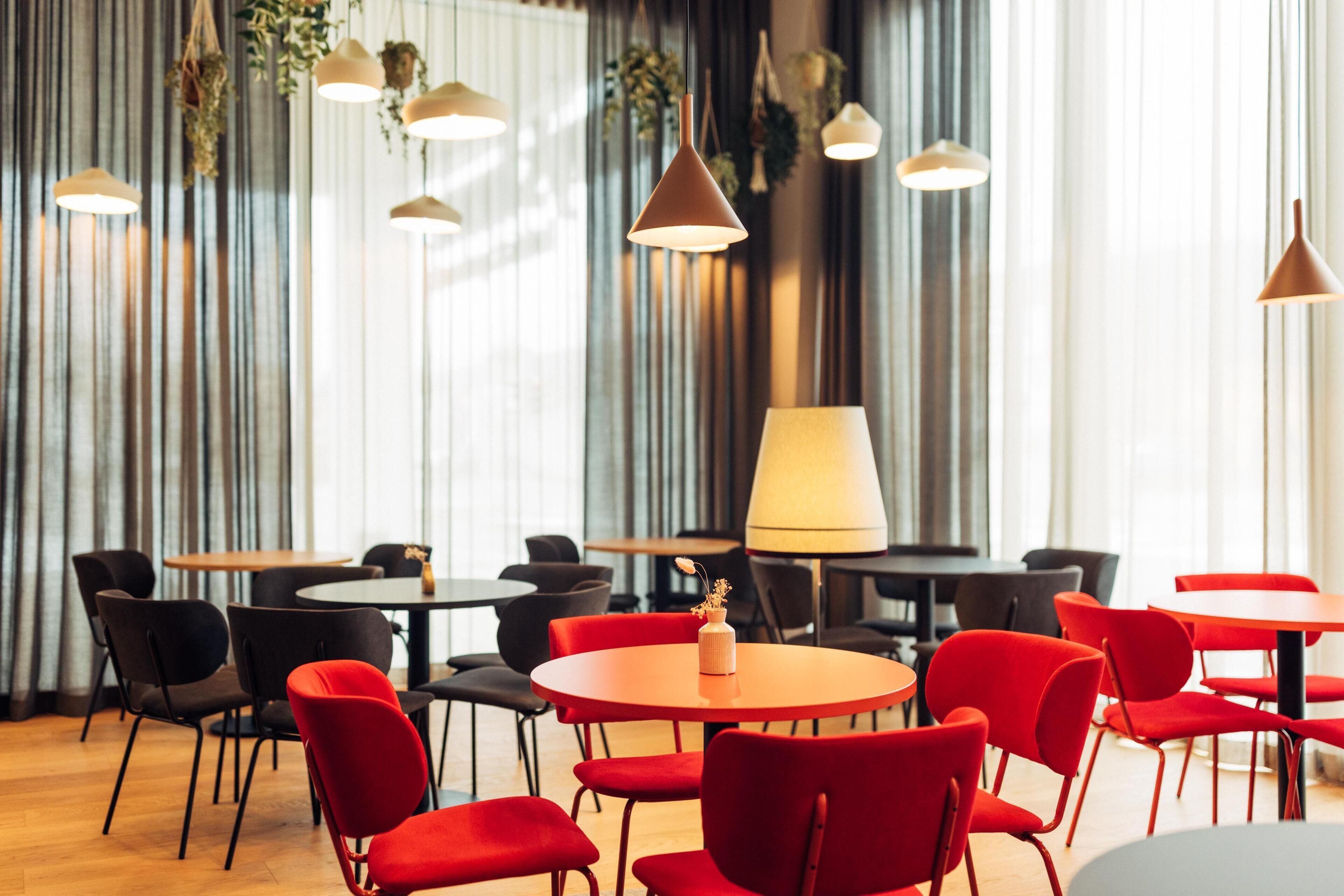 A modern dining area with round tables and red chairs, featuring pendant lighting and a large window with curtains.