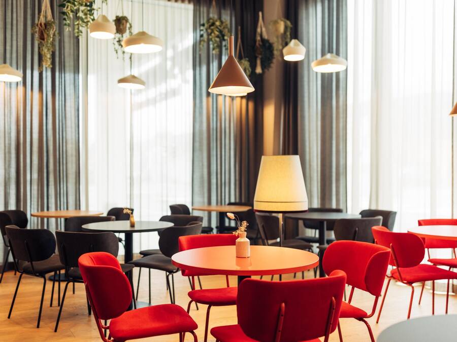 A modern dining area with round tables and red chairs, featuring pendant lighting and a large window with curtains.