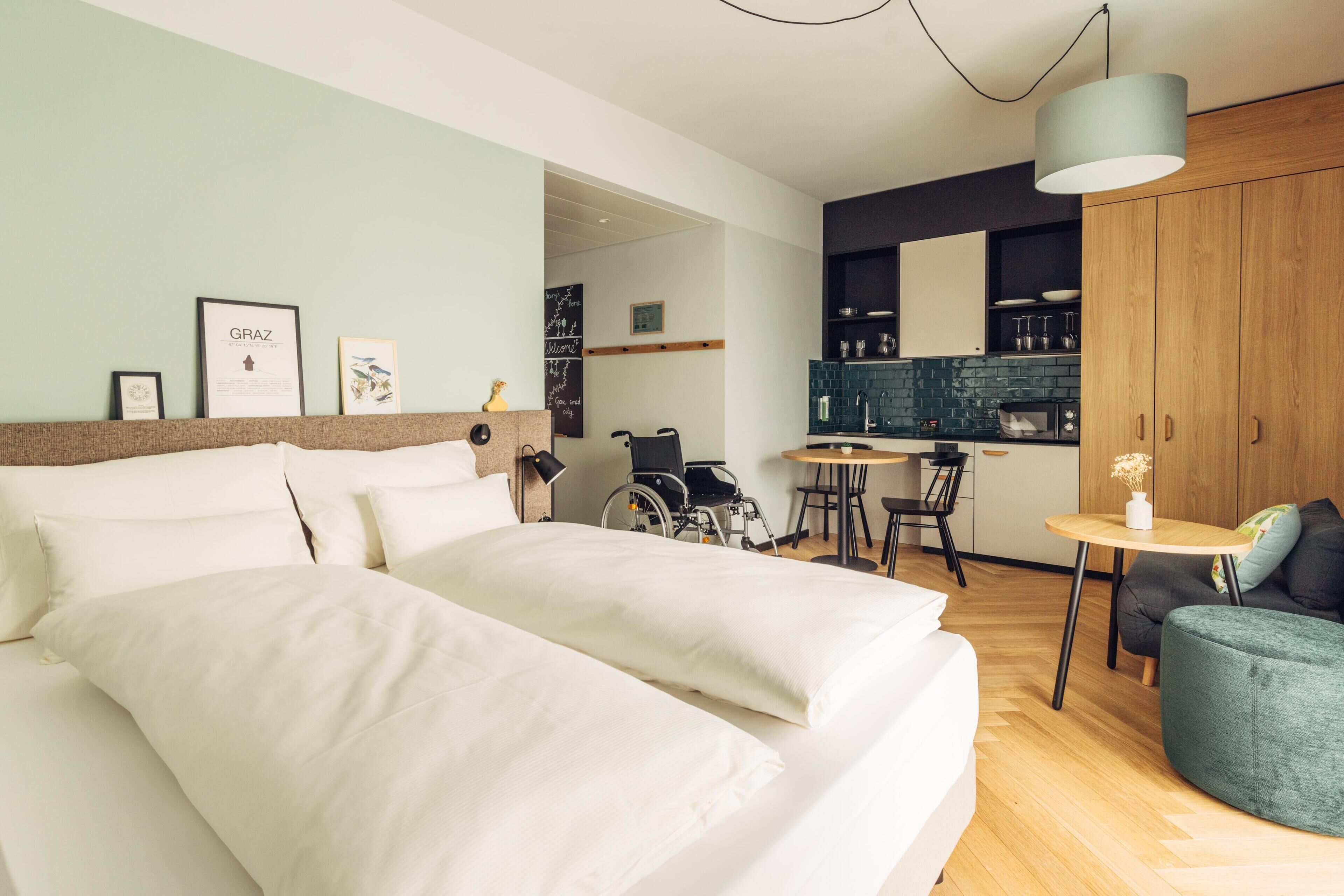 A modern hotel room with a large bed, a wheelchair-accessible chair, a small dining area, and a kitchenette.