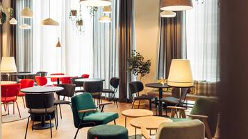A modern hotel lobby with round tables and chairs, large windows with curtains, and hanging pendant lights.