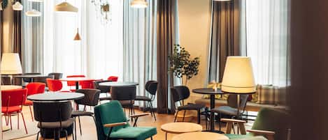 Un moderno vestíbulo de hotel con mesas y sillas redondas, ventanas amplias con cortinas y lámparas colgantes.