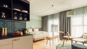 Una habitación de hotel moderna con una cama, un escritorio, una silla, una pequeña mesa, un sofá, una zona de cocina con mostrador y armarios, y un gran ventanal con cortinas.