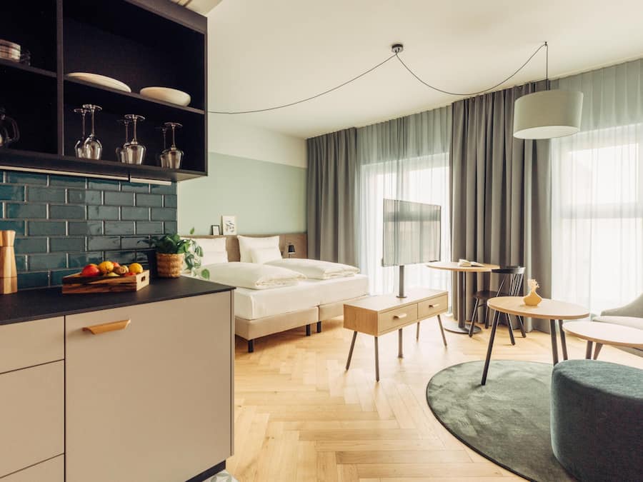 A modern hotel room with a bed, a desk, a chair, a small table, a sofa, a kitchen area with a counter and cabinets, and a large window with curtains.