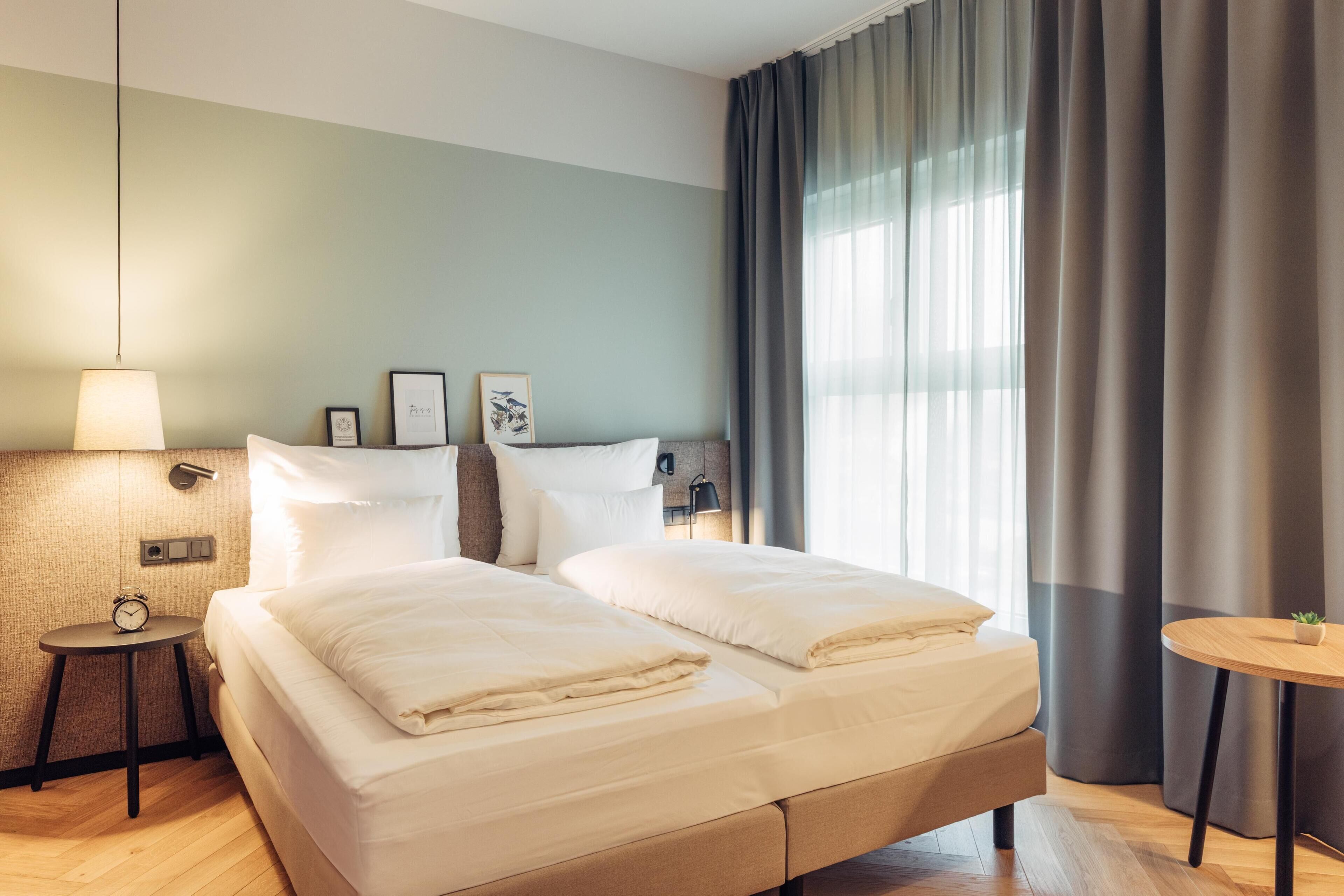 Una habitación de hotel con cama, mesitas de noche, lámpara y ventana con cortinas.
