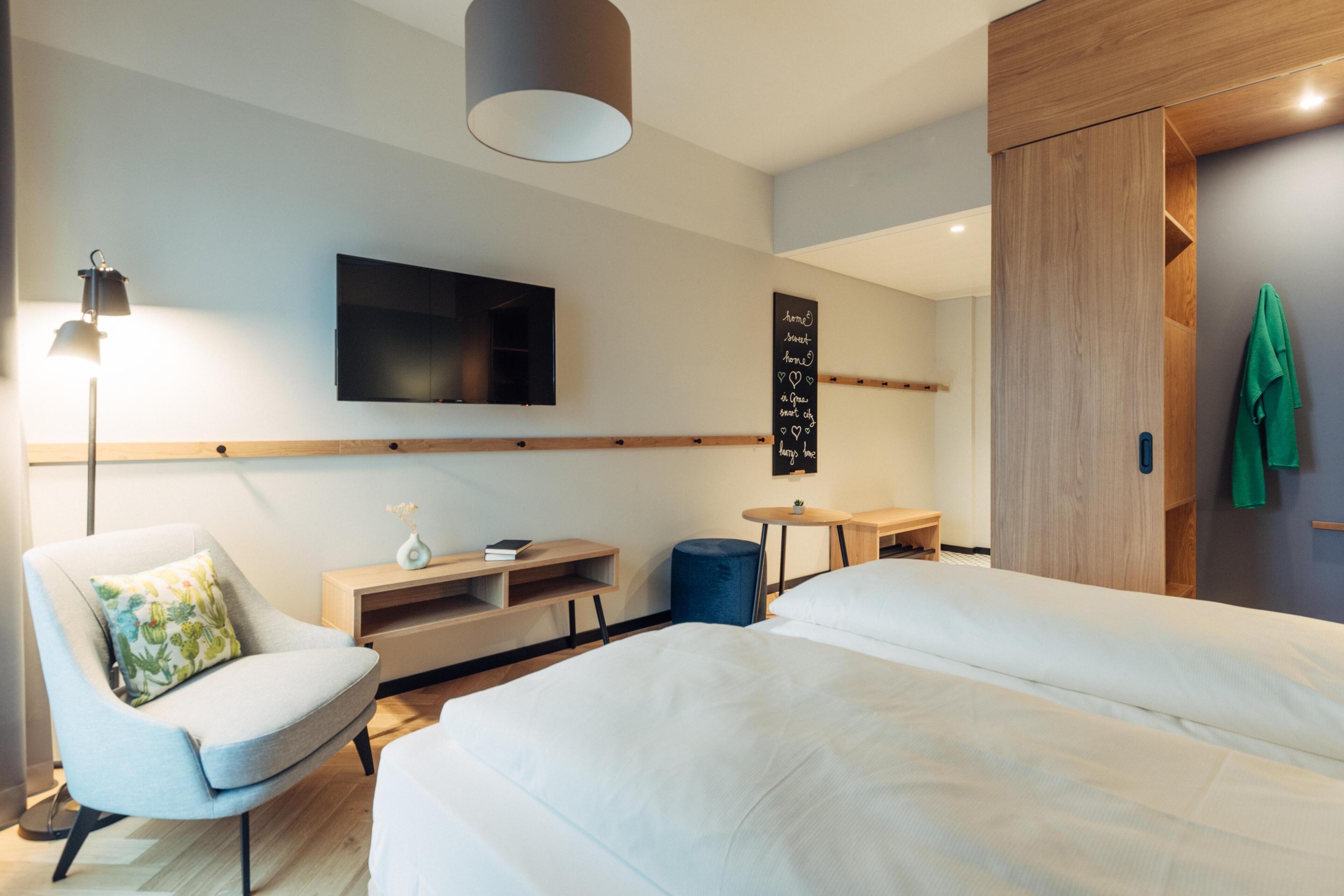 Una habitación de hotel moderna con una cama grande, un televisor de pantalla plana montado en la pared, un escritorio pequeño y una silla con cojín.