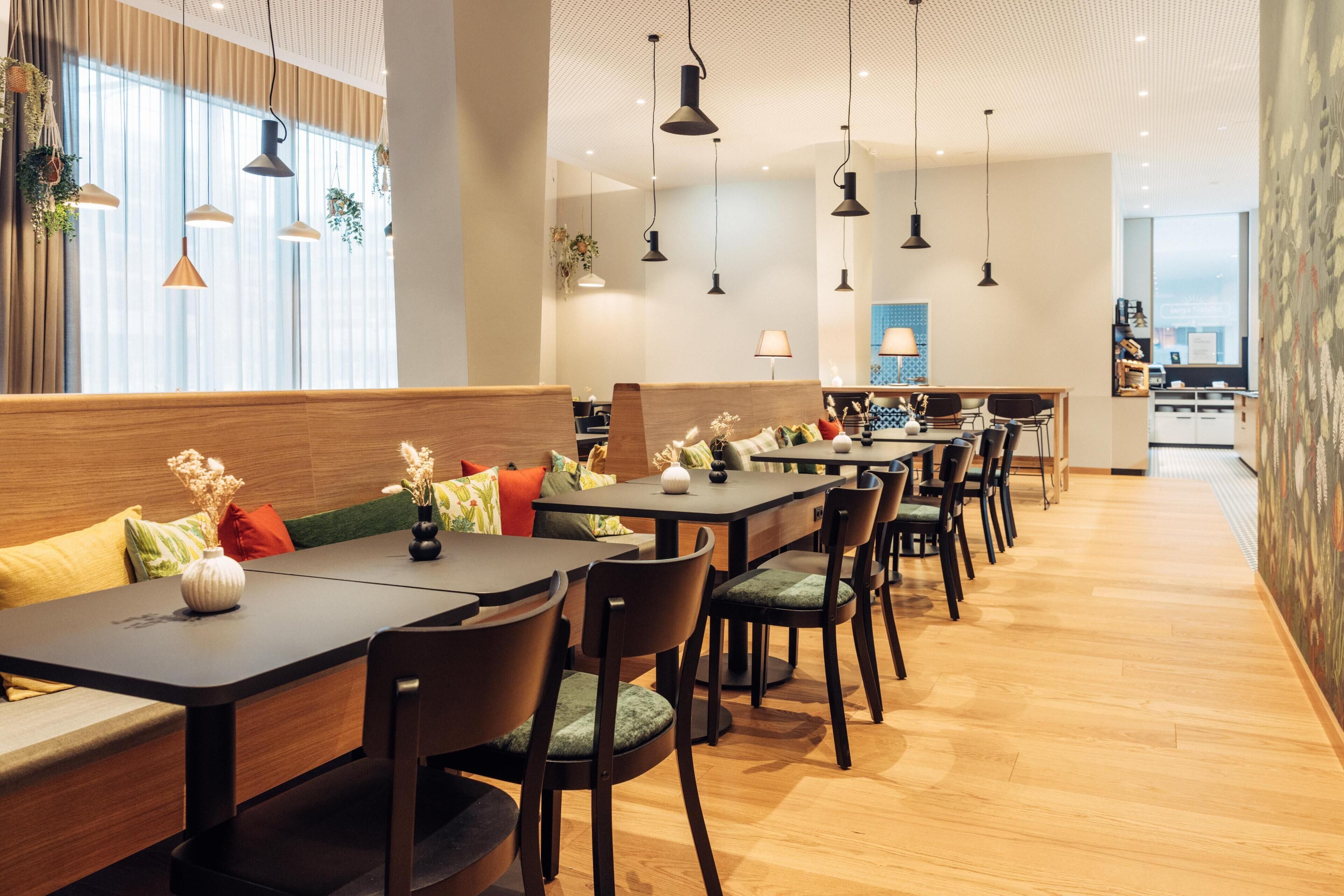 Un restaurante moderno con mesas y sillas de madera, lámparas colgantes y vistas de la ciudad a través de amplios ventanales.