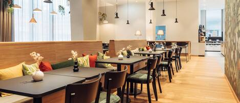 Un restaurante moderno con mesas y sillas de madera, lámparas colgantes y vistas de la ciudad a través de amplios ventanales.