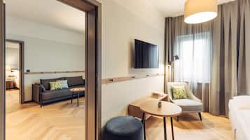 A modern hotel room with a sofa, a round table, a chair, a TV mounted on the wall, and a large window with curtains.