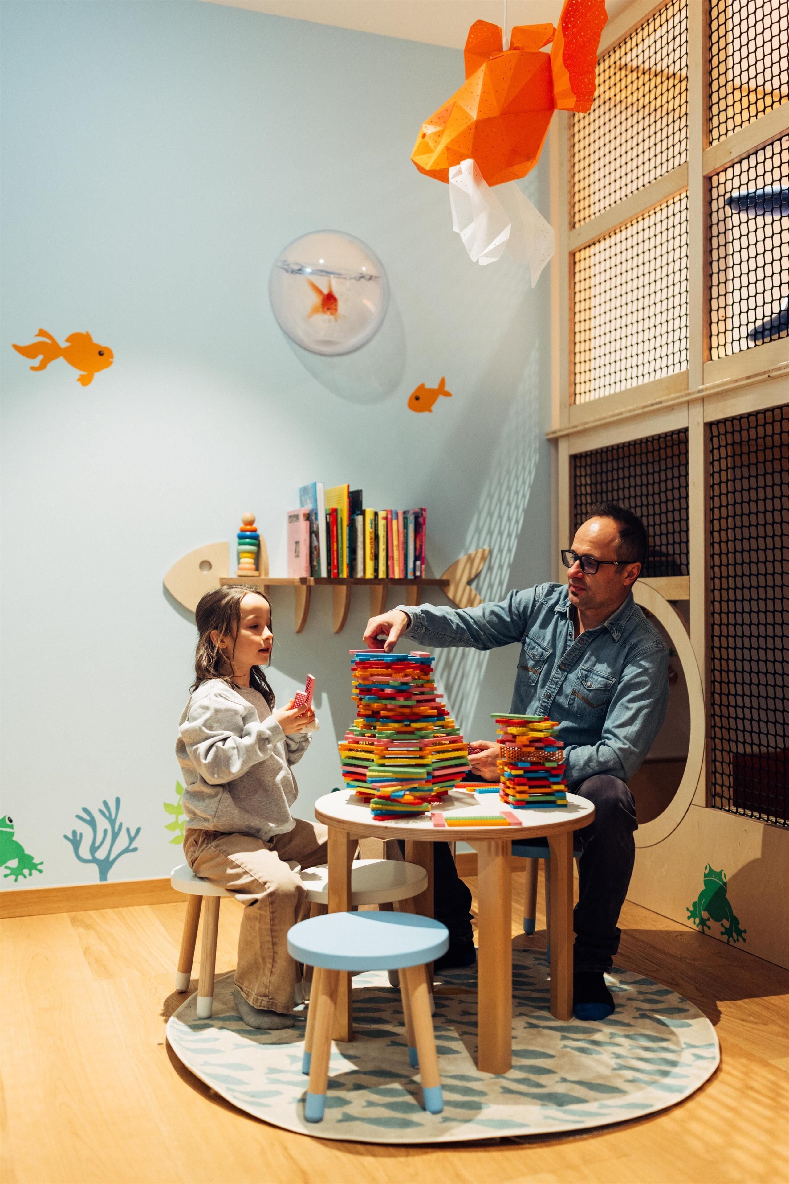 Un niño y un adulto en una mesa redonda de madera con una pila de bloques de colores. La habitación tiene una pared azul claro con un adhesivo de pez, una estantería llena de libros y una ventana con mosquitera.