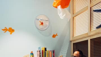 Uma criança e um adulto sentados à mesa redonda de madeira com uma pilha de blocos coloridos. O quarto tem uma parede azul clara com adesivo de peixe, uma estante com livros e uma janela com mosquiteiro.