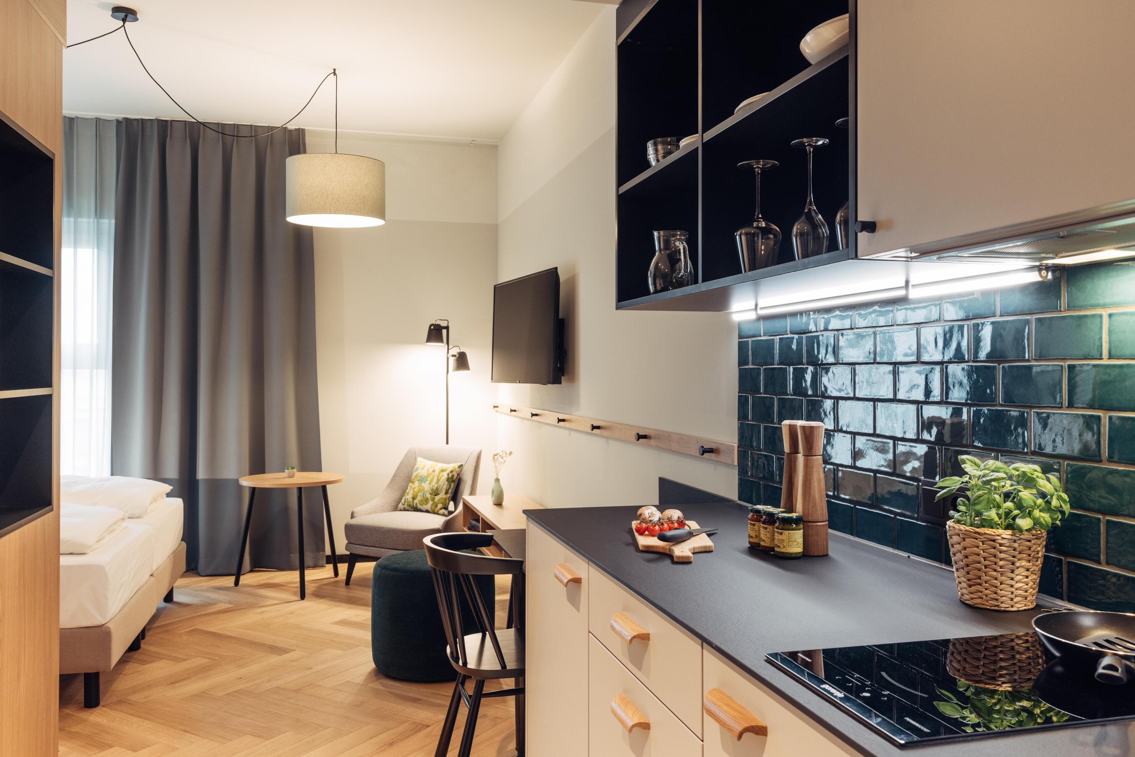 A modern kitchen with a black countertop, wooden cabinets, and a tiled backsplash. There is a dining area with a table and chairs, and a small seating area with a sofa and a lamp.