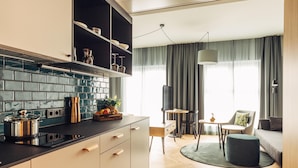 Una cocina moderna con encimera negra, armarios blancos y salpicadero de azulejos. Hay una zona de comedor con una mesa redonda y sillas, y un salón con sofá y lámpara.