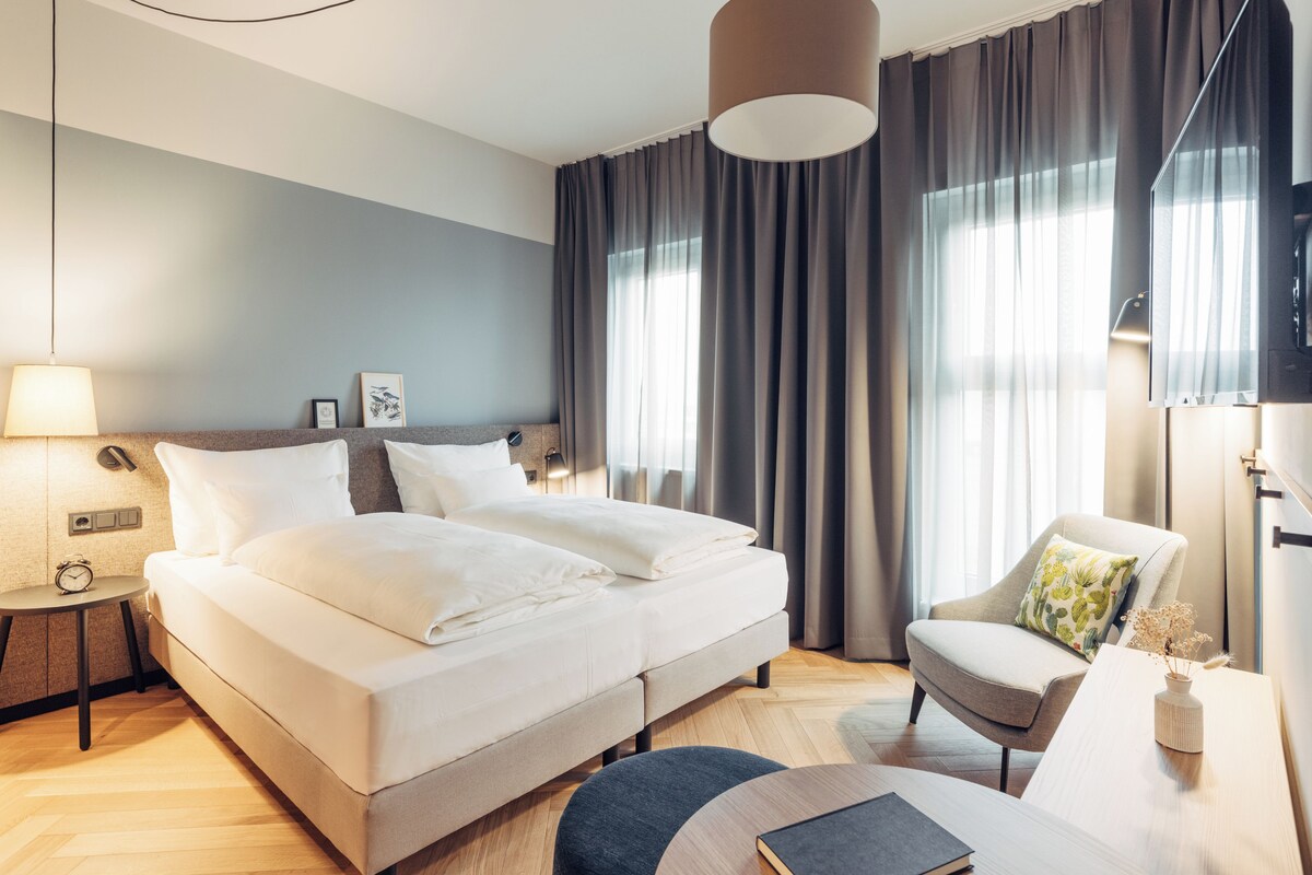 A modern hotel room with a large bed, a side table with a lamp, a chair, and a flat-screen TV.