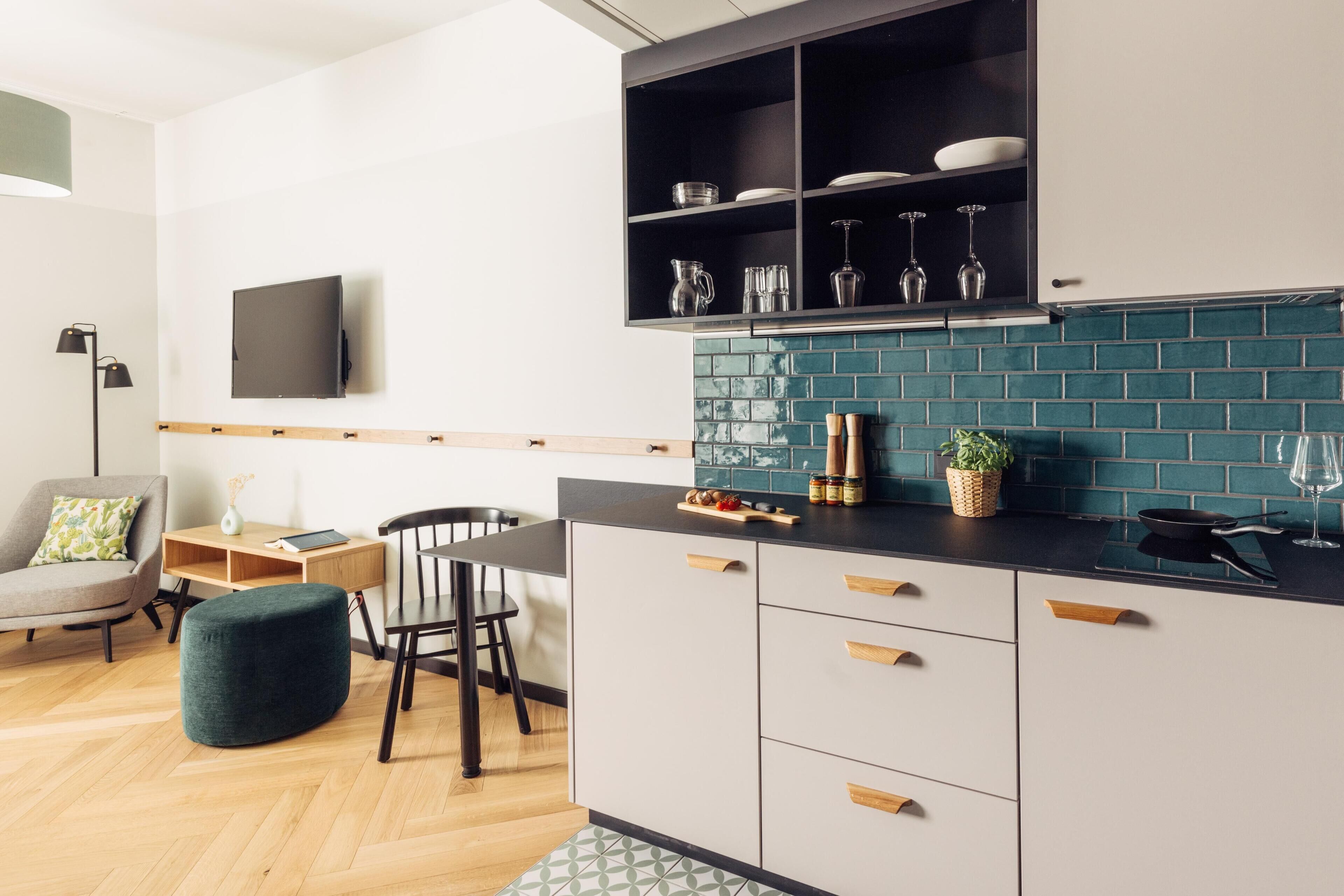 A modern kitchen with a black countertop, white cabinets, and a teal backsplash. There is a dining area with a wooden table and chairs, and a green ottoman.