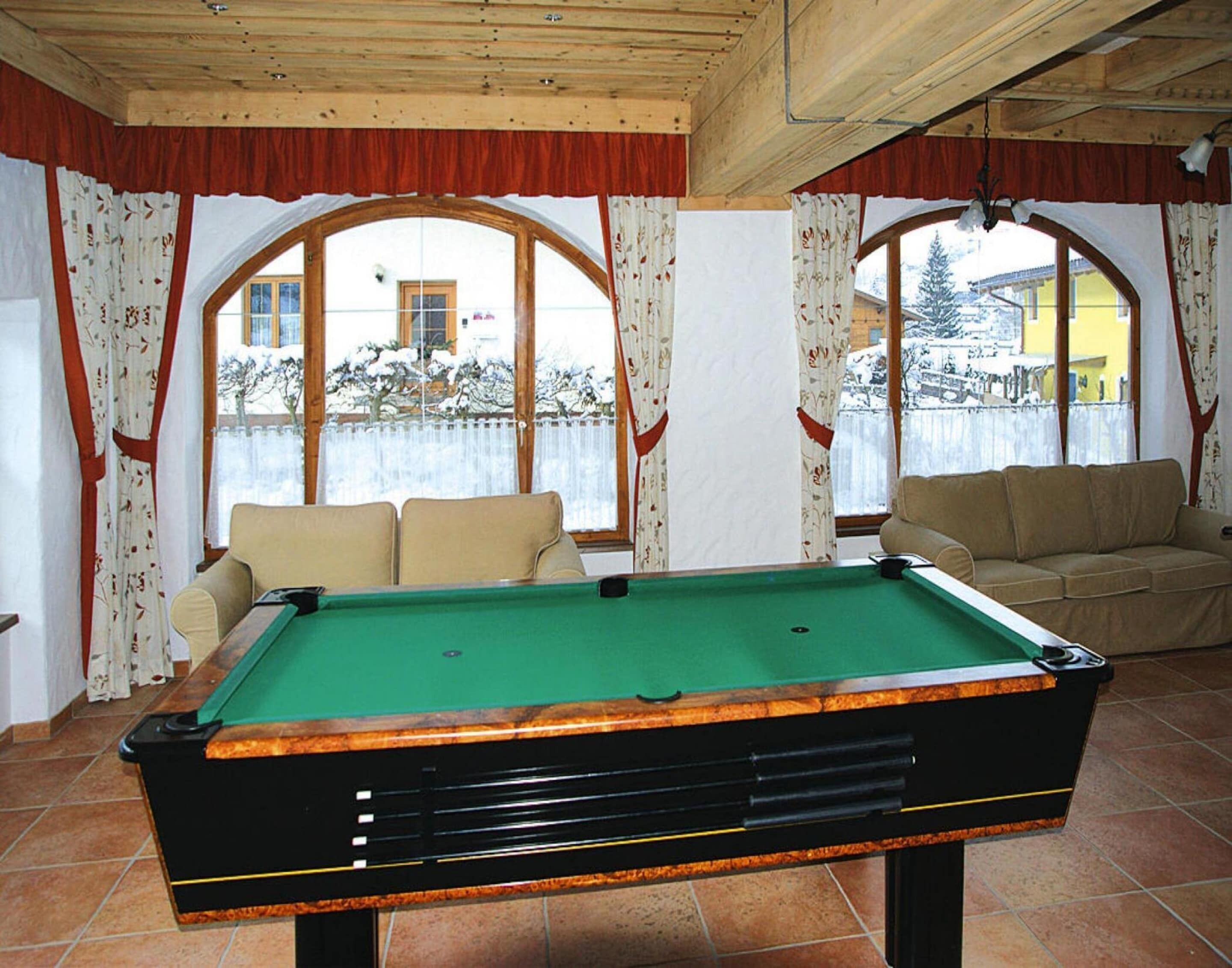 A pool table in a room with two sofas, large windows with curtains, and a view of a snowy landscape.