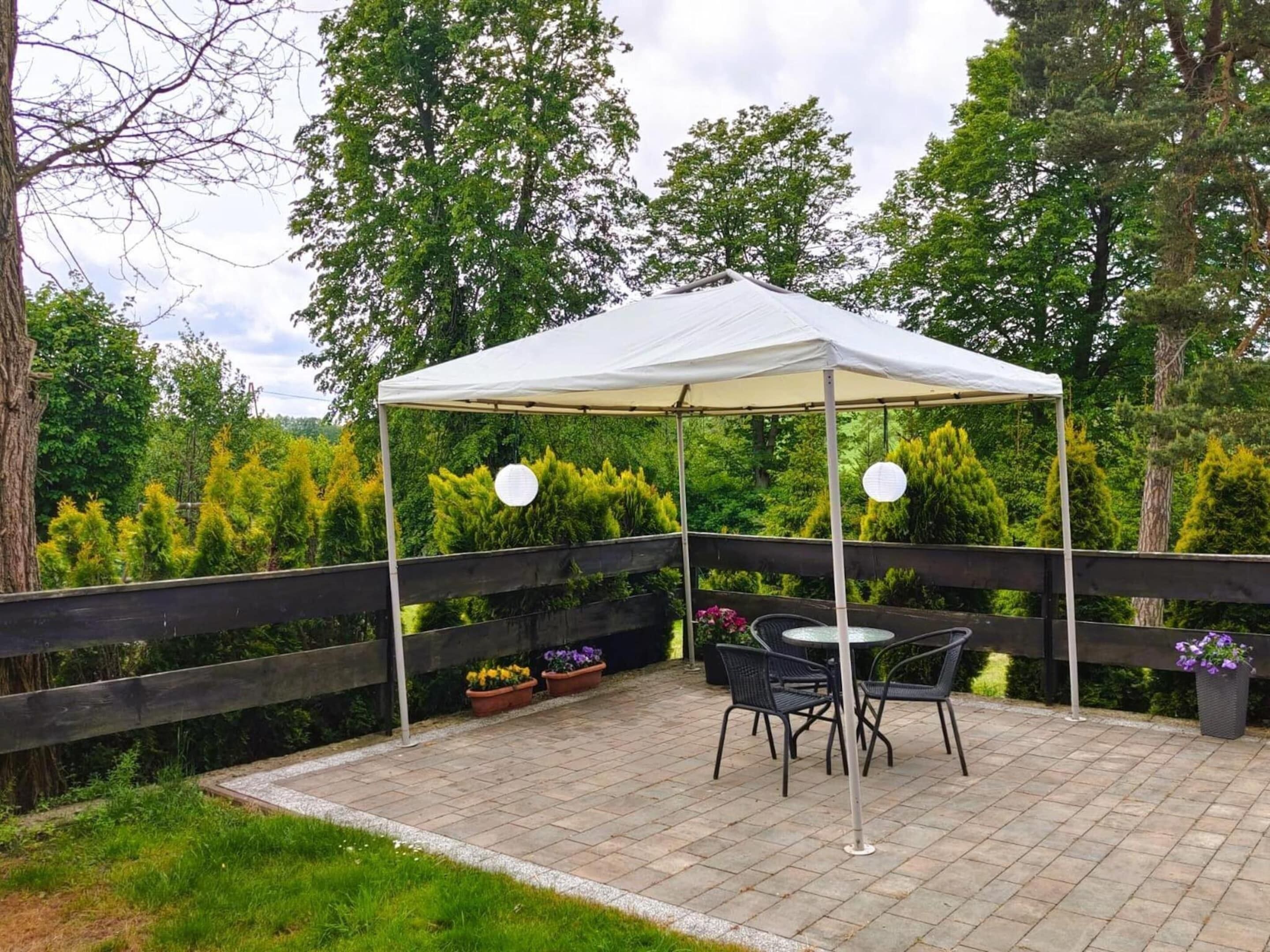 Een overdekt terras met een tafel en stoelen, omringd door een houten omheining en weelderige begroeiing.