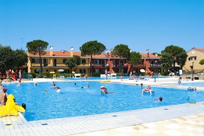 Une zone avec piscine où plusieurs personnes nagent et s’amusent, entourée de bâtiments aux toits recouverts de tuiles rouges et d’arbres.
