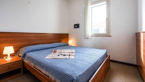 Une chambre à coucher avec une tête de lit en bois, un lit simple recouvert d’une courtepointe à motifs bleus, une table de chevet avec une lampe et une fenêtre avec des rideaux.