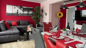 Ein modernes Wohnzimmer mit roter Akzentwand, einem grauen Sofa und einem Essbereich mit roten Stühlen und einem weißen Tisch.