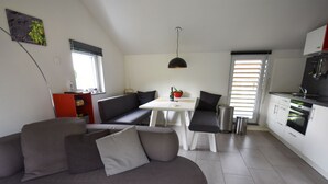 Un salon moderne avec un canapé gris, une table à manger blanche et un coin cuisine doté de meubles blancs et d’un four noir.