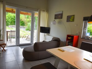 A modern living room with a sofa, a dining table, a TV, and a sliding glass door leading to an outdoor deck. - Holiday Home in Vulkaneifel (Berlingen)