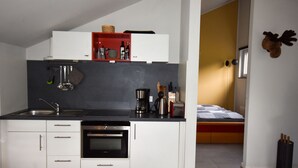 Une cuisine compacte équipée d’un four encastré, d’une machine à café et d’un grille-pain. On y trouve une petite étagère avec des ustensiles de cuisine et un panier. La cuisine donne sur une chambre à coucher qui comprend un lit et une décoration murale représentant une tête de cerf.