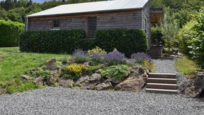 Un chalet rustique en bois, surmonté d’un toit en métal, entouré d’un jardin bien entretenu où poussent diverses plantes, et un chemin de gravier mène à l’entrée.