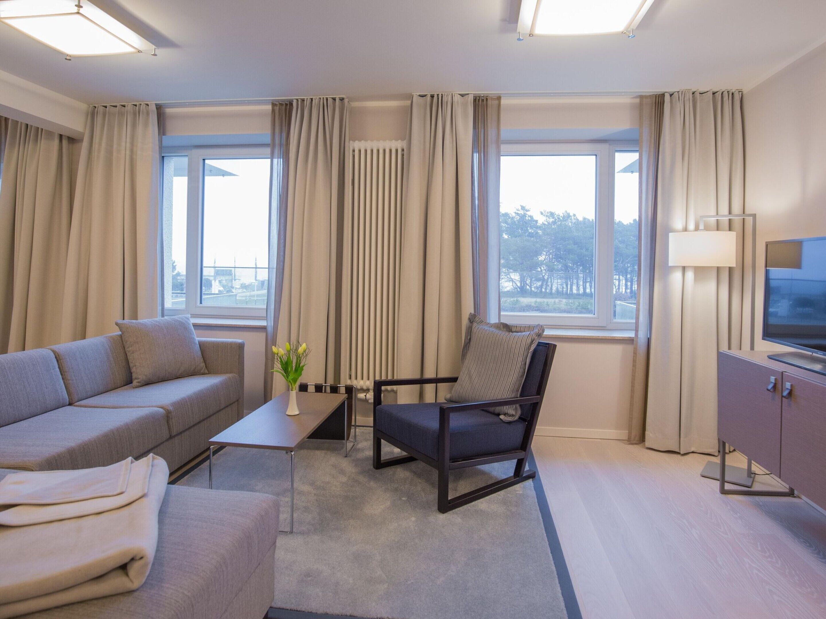 Une chambre d’hôtel moderne équipée d’un canapé, d’un fauteuil, d’une télévision et de grandes fenêtres avec des rideaux.