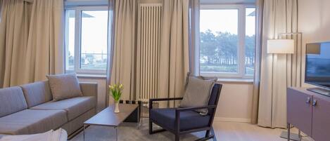 Une chambre d’hôtel moderne équipée d’un canapé, d’un fauteuil, d’une télévision et de grandes fenêtres avec des rideaux.