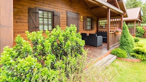 A wooden cabin with a porch, two windows with shutters, and a small garden area with bushes and a potted plant. - Holiday Homes With Terraces, Large Garden (Ustronie Morskie)