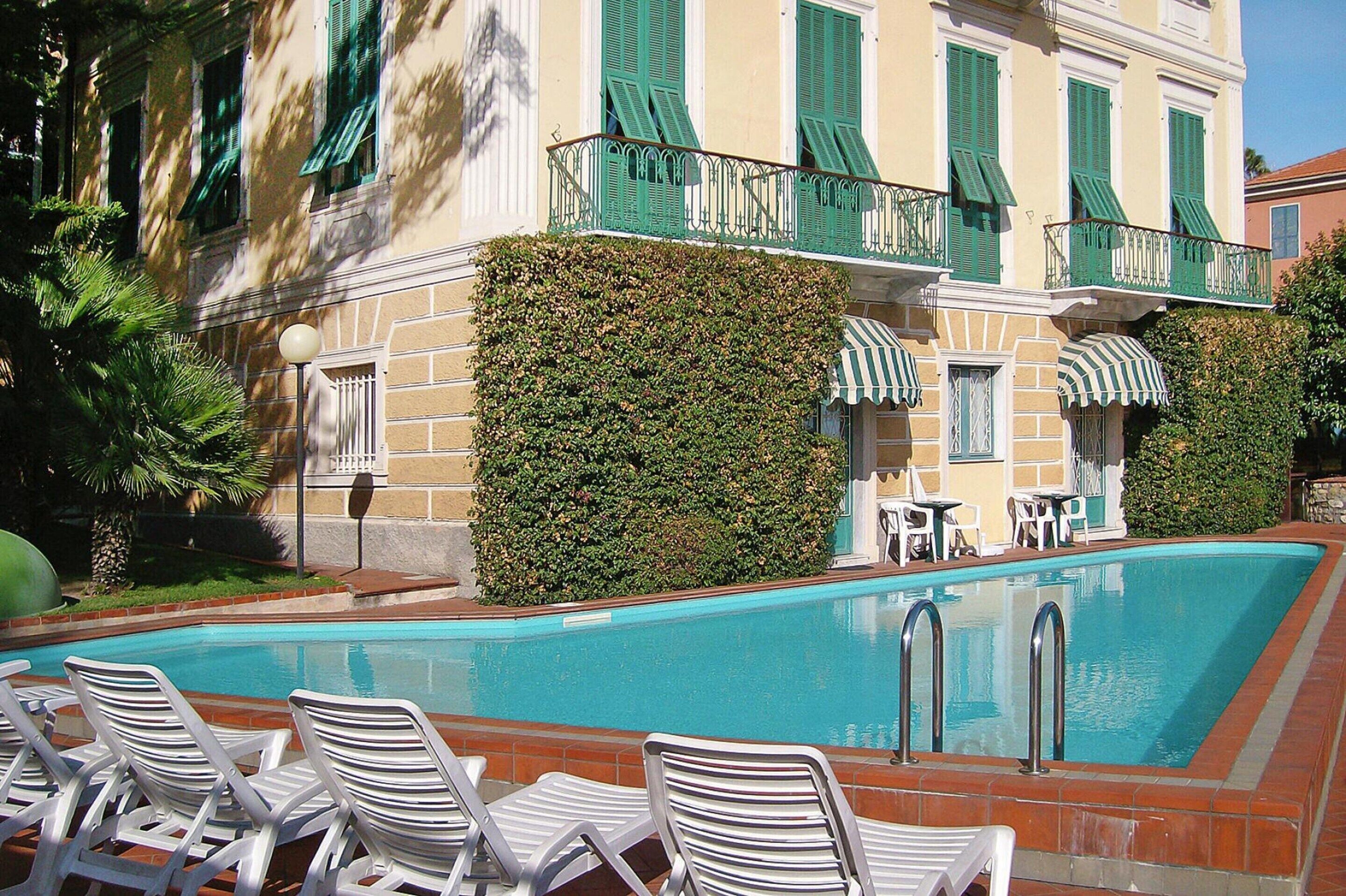 Una piscina con sillones de descanso, un edificio con contraventanas verdes y un seto.