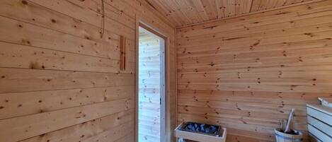 A wooden sauna room with benches, a bench with a bucket, and a door leading outside.