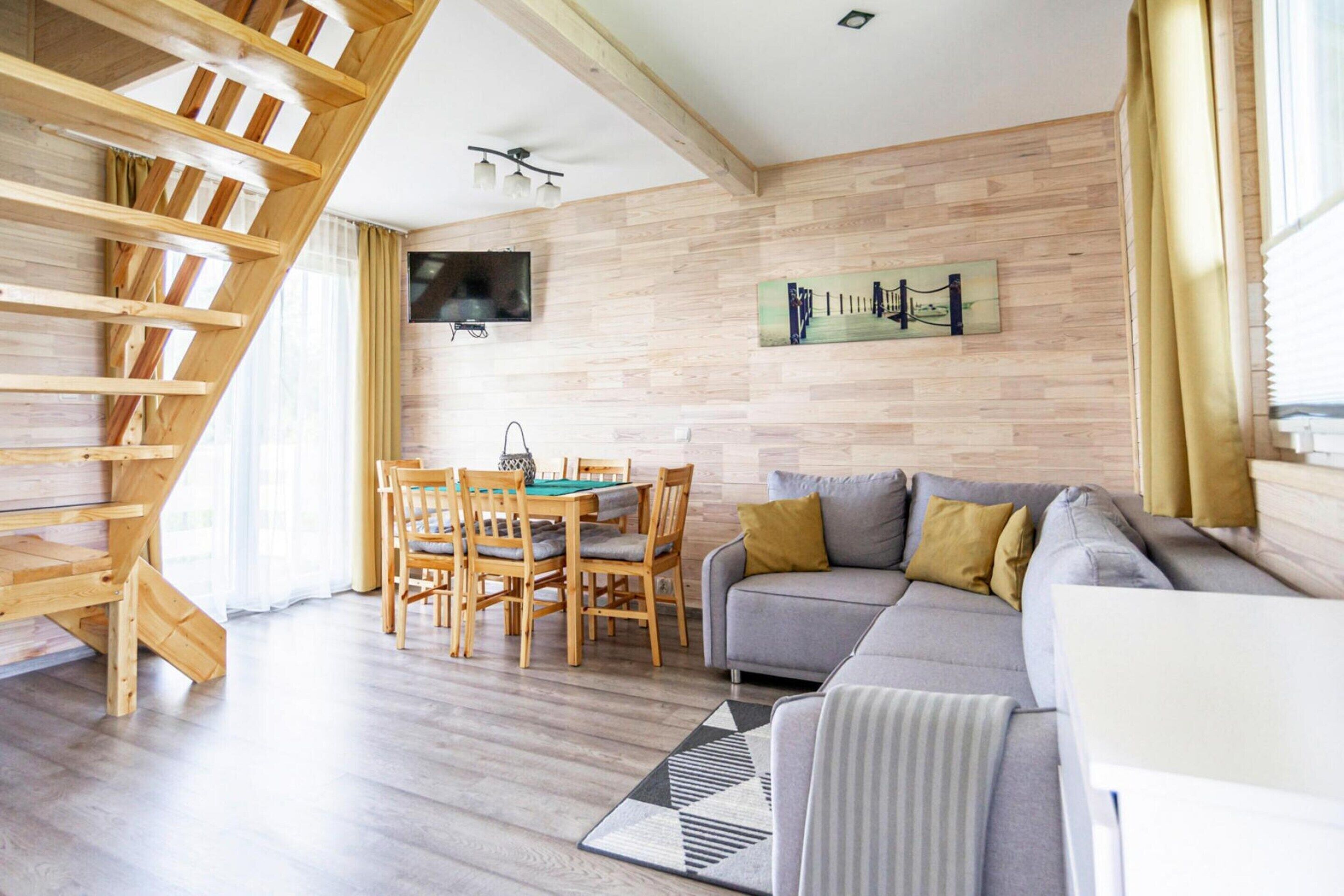 A modern living room with a wooden staircase, a grey sofa, a dining table with chairs, and a flat-screen TV mounted on the wall.