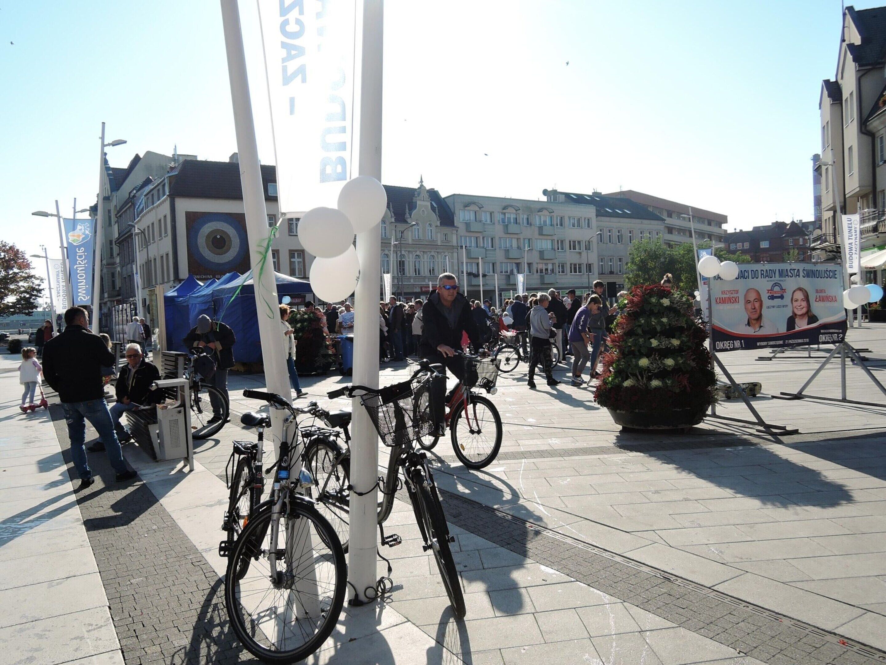 Een openbaar plein met geparkeerde fietsen, een groot bloemstuk en een menigte mensen.