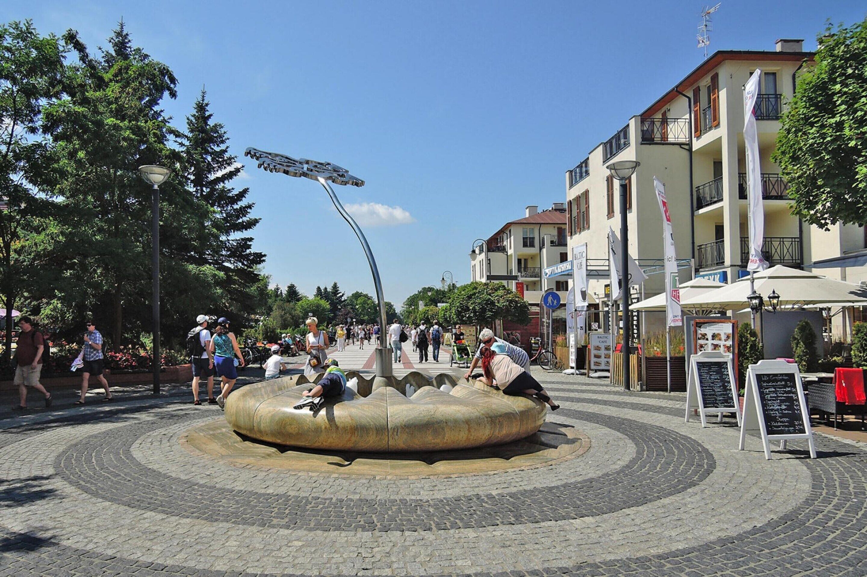 A public square with a circular stone fountain, people walking around, and buildings with balconies and flags.