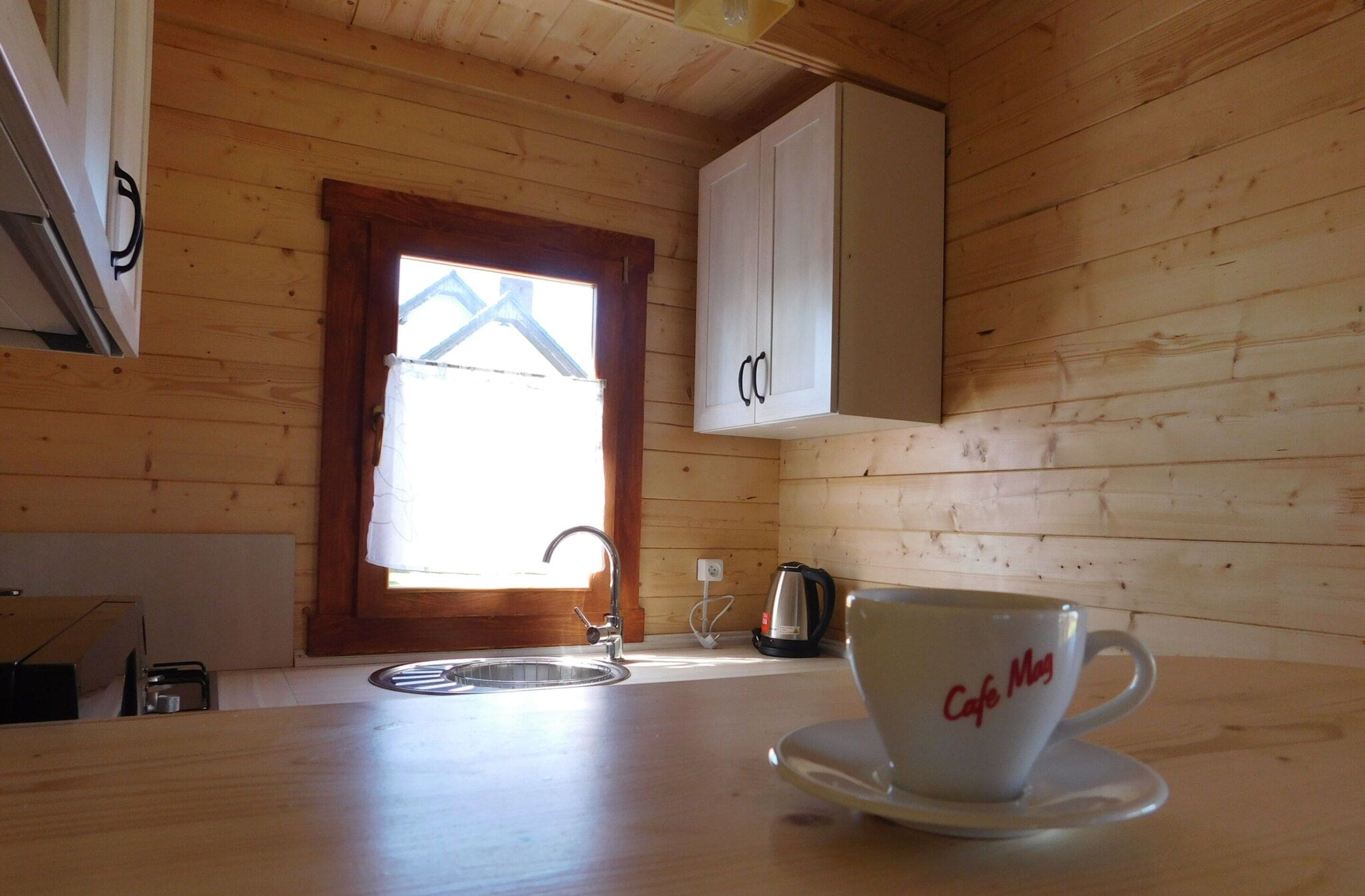 A wooden cabin interior with a kitchen area, including a sink, a kettle, and a cup labeled "Cafe Mug" on a table.