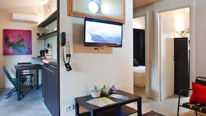 A modern living room with a flat-screen TV mounted on the wall, a dining area with a table and chairs, and a kitchen area with a countertop and cabinets.