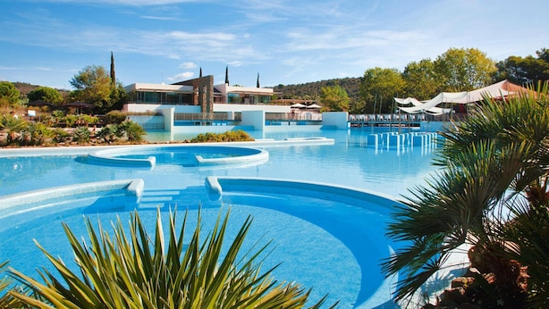 A modern hotel with a large swimming pool, surrounded by lush greenery and a clear blue sky.