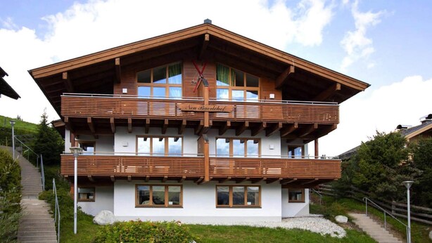 Een houten chalet met twee verdiepingen, met een balkon op de bovenverdieping, gelegen op een helling met een trap erheen.