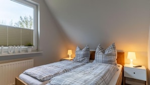 A bedroom with two beds, a window with blinds, a bedside table with a lamp, and a radiator. - Ferienwohnung Strandperle (St. Peter-Ording)