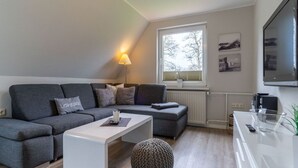 A modern living room with a grey sectional sofa, a white coffee table, a round ottoman, a floor lamp, a large window with blinds, and a flat-screen TV mounted on the wall. - Ferienwohnung Strandperle (St. Peter-Ording)