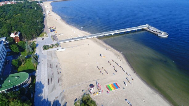 An aerial view of a beach with a long pier extending into the water, a sandy beach with people and umbrellas, and buildings along the shore. - Cozy Holiday Studio, Close to the Beach (Kołobrzeg)
