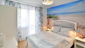 A bedroom with a bed, curtains, a nightstand, a lamp, and a beach mural on the wall. - Cozy Holiday Studio, Close to the Beach (Kołobrzeg)